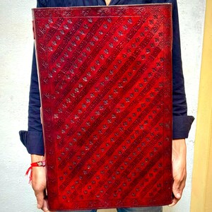 Pode incluir: Um grande item retangular de couro vermelho com um padr&atilde;o de listras diagonais. O couro tem uma apar&ecirc;ncia texturizada com detalhes em relevo. O item &eacute; segurado por uma pessoa vestindo uma camisa azul escura e jeans.