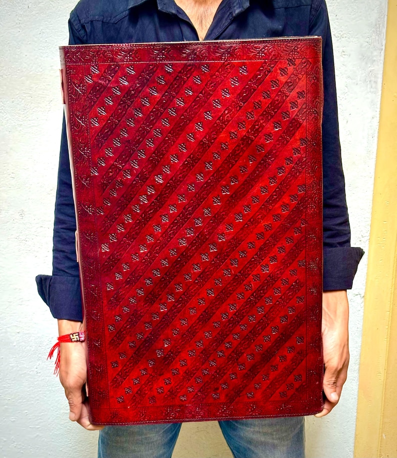 Pode incluir: Um grande item retangular de couro vermelho com um padr&atilde;o de listras diagonais. O couro tem uma apar&ecirc;ncia texturizada com detalhes em relevo. O item &eacute; segurado por uma pessoa vestindo uma camisa azul escura e jeans.
