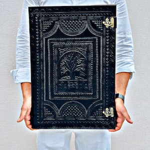 May include: Black leather journal with intricate embossed tree design and decorative border. The journal has a rectangular shape and is secured with two brass-colored clasps. The cover features a detailed, textured pattern.