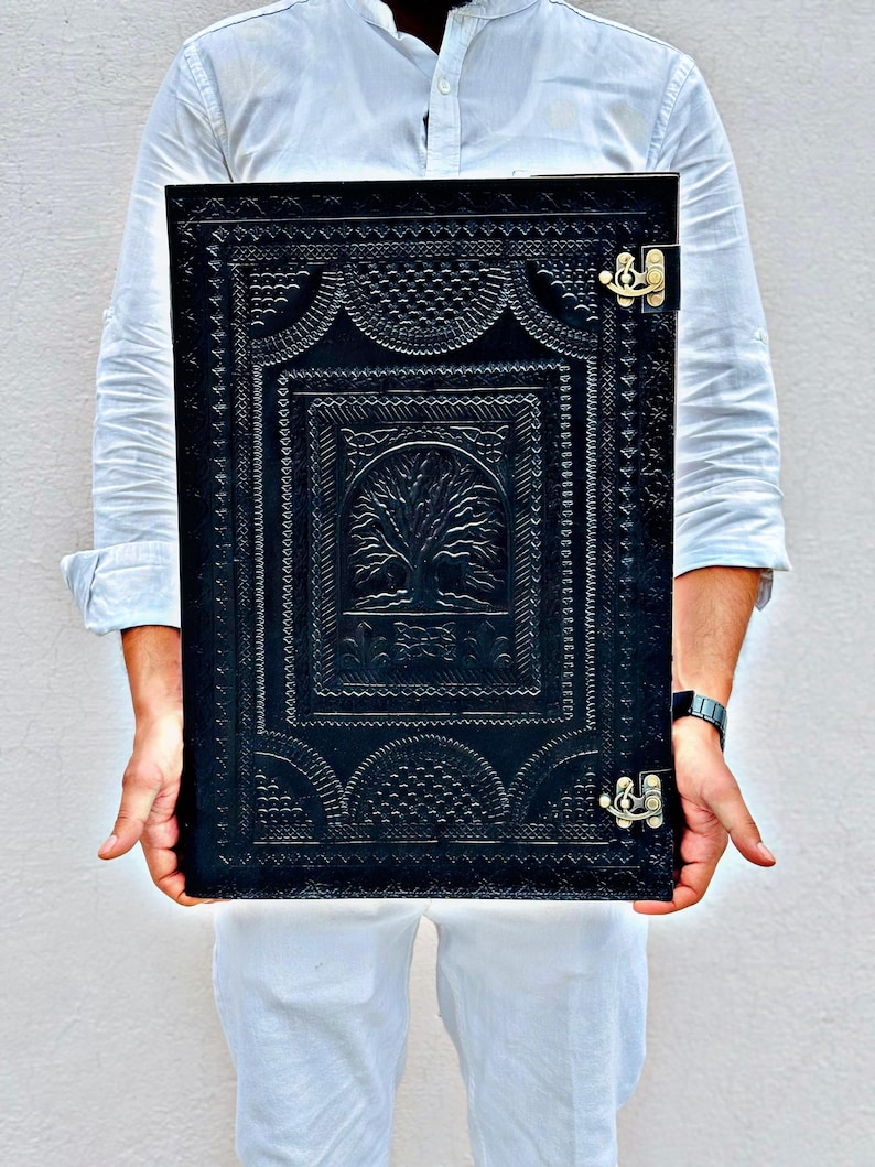 Pode incluir: Di&aacute;rio de couro preto com um intrincado design de &aacute;rvore em relevo e borda decorativa. O di&aacute;rio tem formato retangular e &eacute; preso com dois fechos dourados. A capa apresenta um padr&atilde;o detalhado e texturizado.