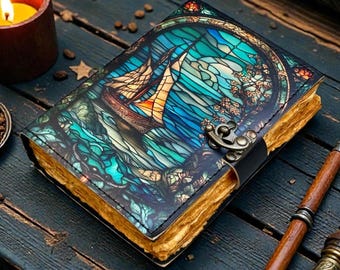 Op de afbeelding: Een leren notitieboek met een glas-in-lood ontwerp dat een zeilboot op de oceaan afbeeldt. De omslag heeft tinten blauw, groen en goud. Het notitieboek heeft een sluiting en verouderde papieren pagina's. Een brandende kaars en pennen liggen in de buurt.