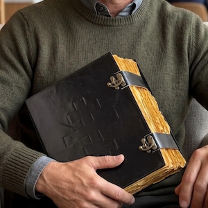 May include: A black leather-bound book with a cross design embossed on the cover. The book has aged, yellowed pages and is secured with two leather straps and ornate metal clasps. The book is held by a person wearing a green sweater.