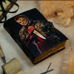 May include: A black leather-bound journal with a graphic of Michael Myers from the Halloween franchise. The journal has a metal clasp and a yellowed paper interior.