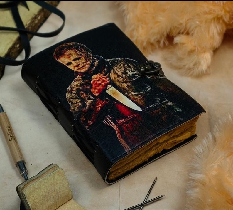 May include: A black leather-bound journal with a graphic of Michael Myers from the Halloween franchise. The journal has a metal clasp and a yellowed paper interior.