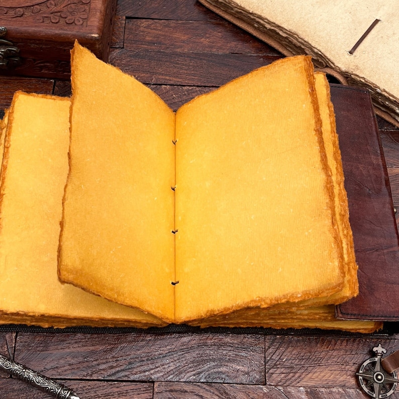 May include: A brown leather-bound journal with yellowed pages is open to reveal blank pages. The journal is lying on a wooden surface.