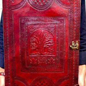 Pode incluir: Um grande di&aacute;rio retangular de couro vermelho com intrincados desenhos em relevo. A capa apresenta um motivo de &aacute;rvore da vida e padr&otilde;es decorativos, fixados por dois fechos de lat&atilde;o. O di&aacute;rio &eacute; segurado por uma pessoa.