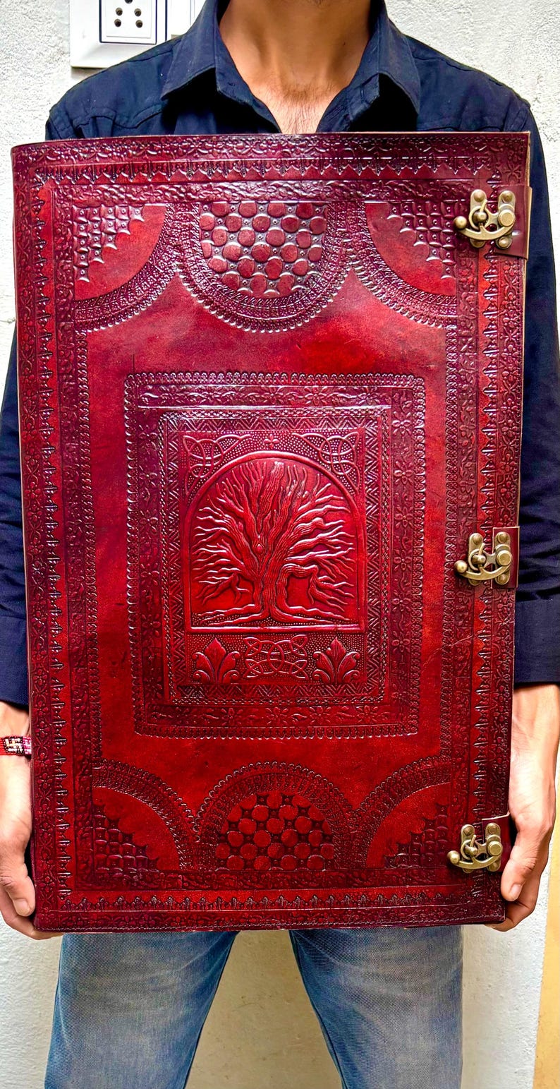 Pode incluir: Um grande di&aacute;rio retangular de couro vermelho com intrincados desenhos em relevo. A capa apresenta um motivo de &aacute;rvore da vida e padr&otilde;es decorativos, fixados por dois fechos de lat&atilde;o. O di&aacute;rio &eacute; segurado por uma pessoa.