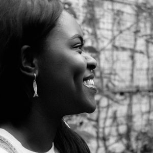 May include: A black and white image of a person smiling, wearing a fish-shaped earring. The earring is a small, dangling hoop with a textured fish design. The person is looking to the side, with a blurred background of foliage.