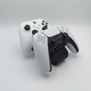 Può includere: Due controller per videogiochi bianchi, uno per Xbox e uno per Playstation, sono posizionati su una stazione di ricarica. La stazione è nera e ha una base bianca.