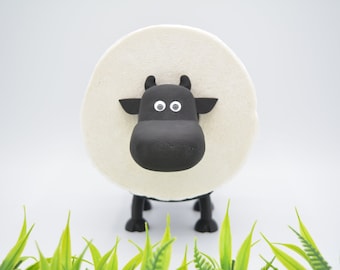 May include: A black and white cow-shaped toilet paper holder with googly eyes. The holder is made of plastic and has a white toilet paper roll attached to it. The holder is sitting on a green grass surface.