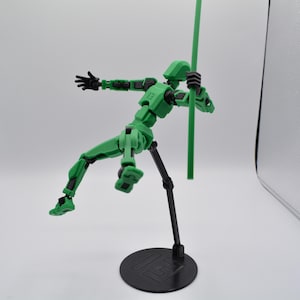 Könnte beinhalten: Eine grüne, artikulierte Actionfigur mit schwarzen Gelenken, die eine grüne Stange hält. Die Figur ist in einer dynamischen Actionpose aufgestellt und steht auf einem schwarzen Sockel mit einem Logo.