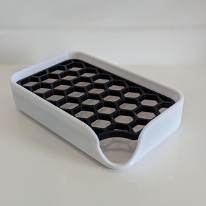 Könnte beinhalten: Ein weißer Seifenschale mit einem schwarzen, wabenförmigen Abtropfgitter. Die Schale hat eine gebogene Vorderkante, um das Abtropfen von Wasser zu erleichtern.