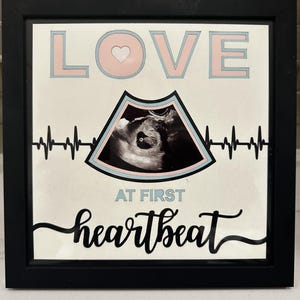 Op de afbeelding: Een ingelijste print met de woorden "LOVE" en "heartbeat" in roze en zwarte letters. De print bevat een echografiebeeld en een hartslaglijn, met de woorden "AT FIRST" in lichtblauw.