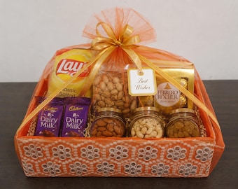 May include: An orange gift basket wrapped in orange tulle with a matching ribbon and a tag reading Best Wishes. It contains a bag of Lay's Classic Salted chips, two Cadbury Dairy Milk bars, a box of Ferrero Rocher, and jars of nuts and raisins.