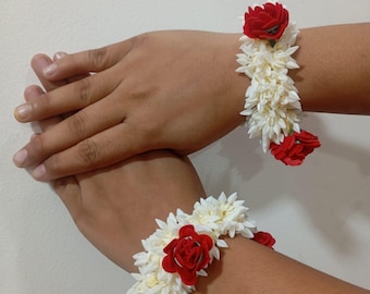 May include: Two floral wristbands, each featuring white flower clusters and a central red rose. The wristbands are worn on wrists, showcasing the contrast of colors and textures. The background is a plain, light-colored wall.