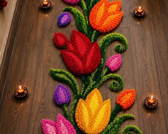 May include: A floral rangoli design crafted from colorful pom-poms in shades of pink, red, orange, yellow, and purple, featuring green stems and swirling leaves, arranged on a dark wood floor and surrounded by small lit tea light candles.