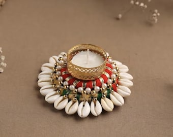 May include: Decorative candle holder featuring a gold-colored metal cup holding a white candle. The base is adorned with white seashells, red and green fabric, and pearl-like beads. The candle holder is on a brown surface.