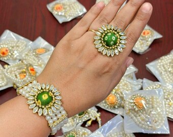 May include: A gold-toned ring and bracelet set featuring a green center stone surrounded by clear, oval-shaped accents. The bracelet and ring are adorned with white pearls. Several similar jewelry pieces are visible in the background.