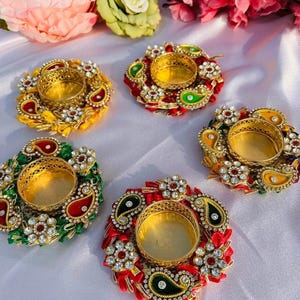 Op de afbeelding: Vijf ronde kaarsenhouders met centrale goudkleurige metalen cups omringd door stoffen ringen. Elke ring is versierd met kleurrijke paisley-vormen, bloemaccenten met heldere strass-steentjes en levendige stoffen bloemblaadjes in geel, rood en groen.