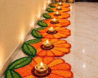 May include: A row of seven circular floral-shaped floor decorations made of orange and yellow petals with green leaf accents. Each flower features a small, metallic gold-toned oil lamp placed in the center, with a lit flame glowing brightly.