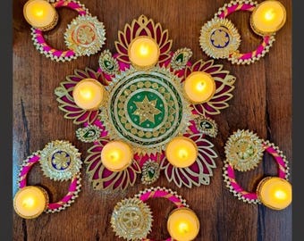 May include: A decorative arrangement featuring lit candles in gold holders, surrounded by ornate, gold-edged floral designs with pink and green accents. The candles are arranged on a dark wood surface, creating a warm, inviting ambiance.
