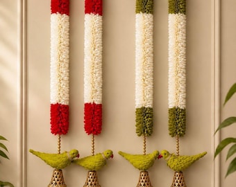 May include: Four vertical wall hangings featuring cylindrical floral garlands. Two garlands are red and white, and two are olive green and white. Each hangs from a gold hook and terminates in a green bird figure above a gold lattice bell.