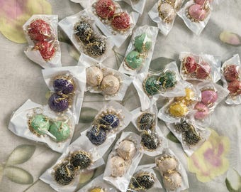 May include: A collection of jhumka earrings in various colors including red, blue, green, pink, yellow, and white, each pair individually packaged in clear plastic. The earrings feature intricate metallic patterns and dangling bell-shaped designs.