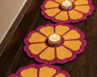 May include: A series of circular, flower-shaped crochet mats arranged in a line. Each mat features a bright yellow center with eight petal-shaped sections and a vibrant magenta border. A small, clear glass tea light candle holder sits in the center.