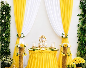 May include: A decorative display featuring a central table draped in a yellow pleated cloth, topped with a white Ganesha statue and two candles. The backdrop consists of layered yellow and white curtains adorned with yellow and white floral garlands.