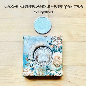 May include: Two silver coins, one above a floral patterned box. The top coin has an intricate design. The bottom coin is set in the box and features a seated figure. Text at the top reads "LAXMI KUBER AND SHREE YANTRA" and "10 Grams".