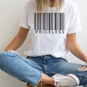 以下が含まれることがあります： 黒いバーコードのデザインと「PRICELESS」の文字がプリントされた白いTシャツ。Tシャツは青いジーンズと白いスニーカーと組み合わせています。人物は座っています。