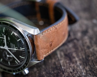 May include: Close-up of a silver wristwatch with a black face and a brown leather strap. The watch face displays white hands and markers, with the brand name and model details visible. The leather strap has a textured appearance and is stitched with a contrasting thread.