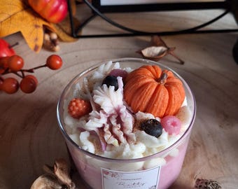 Op de afbeelding: Een decoratieve kaars in een doorzichtige glazen container, met een pompoen- en bessenontwerp. De kaars heeft een roze basis en is bedekt met witte was, een oranje pompoen en donkere bessen. De tekst "Candle LIT WISHES" is zichtbaar op de achtergrond.