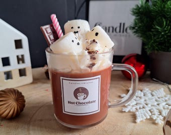 Może przedstawiać: Przezroczysty szklany kubek wypełniony gorącą czekoladą, zwieńczony bitą śmietaną i piankami marshmallow. Różowa i biała słomka w paski oraz baton czekoladowy umieszczone w kubku. Kubek ma etykietę z napisem "Hot Chocolate".