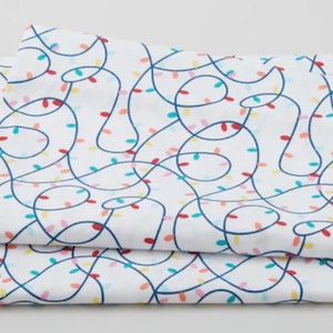 May include: White bed sheets with a repeating pattern of blue string lights with colorful bulbs in red, yellow, teal, and pink. The sheets are folded and stacked, showcasing the design.