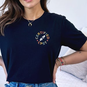 Pode incluir: Uma camiseta preta com um gr&aacute;fico colorido que diz "CERTIFIED CPST" em um c&iacute;rculo com estrelas. A pessoa que est&aacute; usando a camiseta est&aacute; em p&eacute; na frente de uma cama branca com um travesseiro branco e cinza.