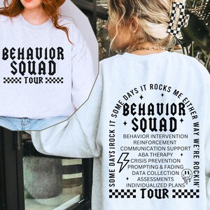 May include: White sweatshirt with black text that reads "Behavior Squad Tour" and a graphic of a lightning bolt and a smiley face. The text also includes a list of services offered by the squad, such as behavior intervention, reinforcement, communication support, ABA therapy, crisis prevention, prompting and fading, data collection, assessments, and individualized plans.