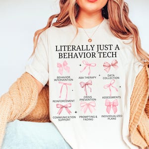 May include: White t-shirt with the text "LITERALLY JUST A BEHAVIOR TECH" and a graphic of pink bows representing different aspects of behaviour therapy, including behaviour intervention, ABA therapy, data collection, reinforcement, crisis prevention, assessments, communication support, prompting & fading, and individualized plans.