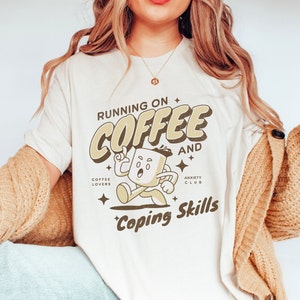 May include: White t-shirt with a brown cartoon coffee cup character running and the text "Running on Coffee and Coping Skills". The text "Coffee Lovers" and "Anxiety Club" are also included.