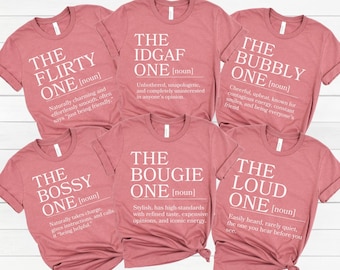 May include: Six dusty rose-colored t-shirts with white text. Each shirt features a different phrase: "The Flirty One," "The IDGAF One," "The Bubbly One," "The Bossy One," "The Bougie One," and "The Loud One." Each phrase is followed by a definition.