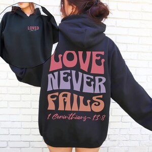 May include: Black hooded sweatshirt with the text "LOVE NEVER FAILS 1 Corinthians 13:8" printed on the back in a colorful retro font. The front of the sweatshirt has the text "LOVED" printed in a smaller font.