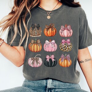 May include: A gray t-shirt with a pattern of nine pumpkins with bows. The pumpkins are in various colors, including orange, pink, and black. Some pumpkins have leopard print bows.