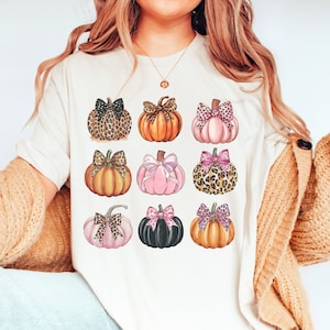May include: A white t-shirt with a design of nine pumpkins with bows. The pumpkins are in various colors, including orange, pink, and black. Some of the pumpkins have leopard print bows.