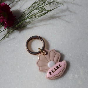 May include: A keychain featuring a pink seashell and a pink oval charm with the word "PEARL" in black lettering. The keychain has a gold-colored clasp. The background is a white marble surface with floral accents.