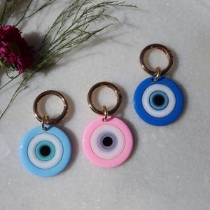 May include: Three round evil eye charms in blue, pink, and blue with gold-colored clasps. Each charm features concentric circles of white, blue, and black. The charms are displayed on a light-colored surface with floral and plant accents.