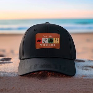 May include: Black baseball cap with a brown patch that says "Live the Wild-Life" with a bear, paw prints, a mountain, and trees on it. The cap is sitting on a sandy beach.