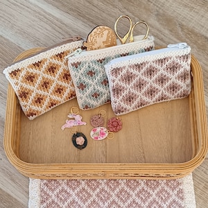 Peut inclure: Trois petites pochettes à fermeture éclair en crochet avec un motif en losange en marron, bleu et rose. Les pochettes sont présentées sur un plateau en bois avec des breloques florales décoratives.