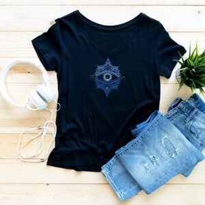 Puede incluir: Una camiseta negra de cuello en V con un diseño de mandala azul y blanco con un ojo en el centro. La camiseta está colocada sobre una superficie de madera con unos auriculares blancos y unos vaqueros azules.