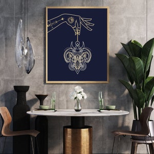 Puede incluir: Un dibujo de línea dorada de una cabeza de carnero con cuernos y una luna creciente por encima, colgando de una mano con un dibujo de línea dorada de un sol y una luna en la muñeca, sobre un fondo azul oscuro.