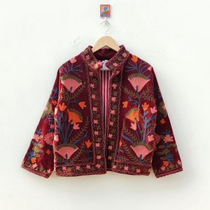 May include: A deep burgundy velvet jacket featuring intricate floral embroidery in shades of pink, orange, and blue. The jacket has a high collar, long sleeves, and a front opening that reveals a red and white striped lining on the interior.